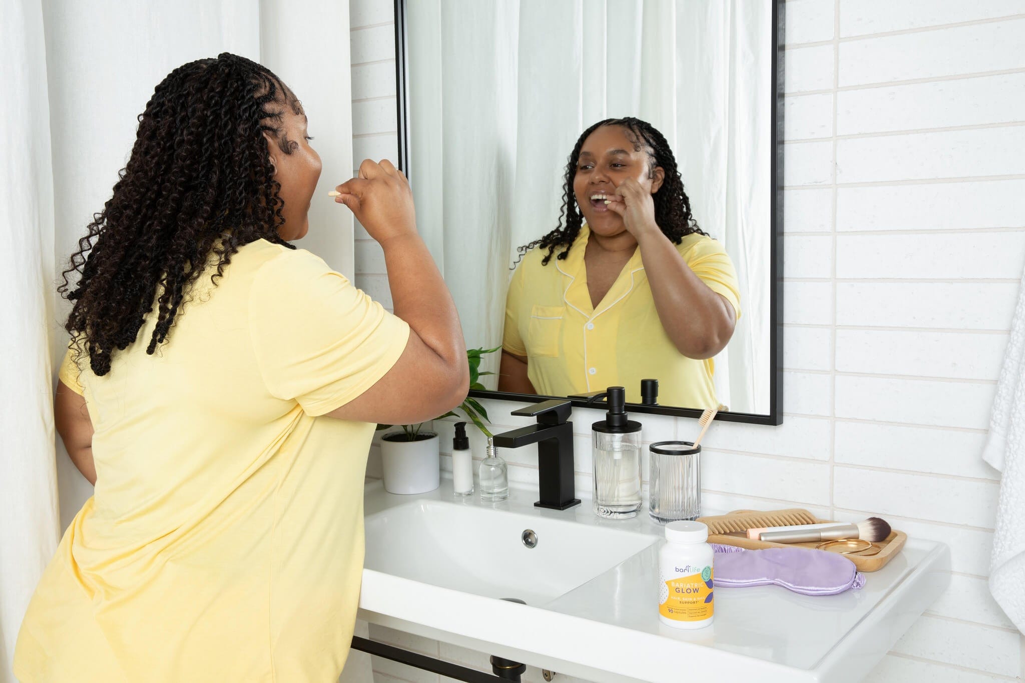 Woman in yellow shirt brushing teeth at bathroom sink, reflecting in mirror with beauty products near.